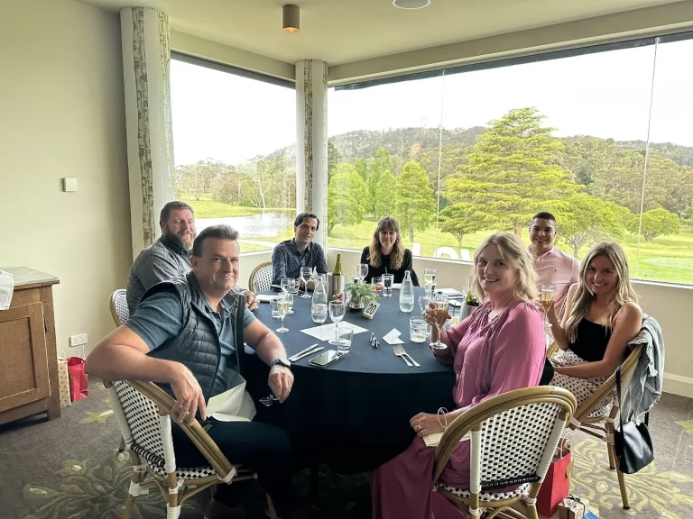 A diverse team from Quattro Architecture enjoying a celebratory lunch in a bright dining room with floor-to-ceiling windows overlooking a park.