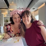 Two women smiling at a Quattro Architecture team event, wearing festive headpieces in a modern interior with white paneled walls and pendant lights.